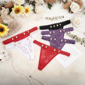 May include: A collection of thong underwear in various colors, including red, white, black, and purple. Each thong features a lace trim and the word "BABY" spelled out in gold letters across the waistband. The underwear is displayed on a soft, white surface.
