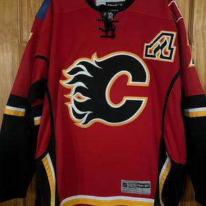 May include: Red hockey jersey with black and yellow accents. Features the Calgary Flames logo on the front, a stylized "C" with flames. The jersey has a lace-up neckline and the number "4" on the shoulder.