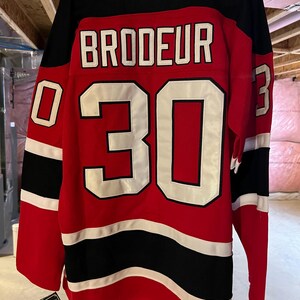 May include: A red, black, and white hockey jersey with the name "BRODEUR" and the number "30" in white block letters. The jersey has a Reebok logo and a tag attached. The jersey is hanging.