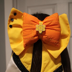 May include: A large, handmade hair bow. The bow has a bright orange centre with yellow stitching and a yellow flower-shaped embellishment. It is set against yellow fabric with black trim and white satin ribbons.
