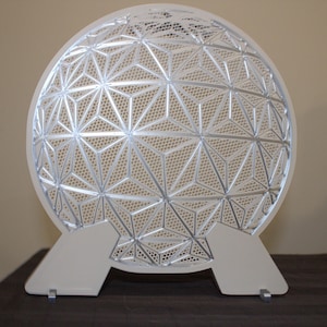 May include: A decorative, geometric, silver-colored sphere with a complex star-like pattern. The sphere is supported by two white, rectangular feet. The design is modern and intricate, suitable for home decor.