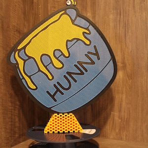 May include: A whimsical honey pot design with the word "HUNNY" in black lettering. The pot is blue with yellow honey dripping down. A yellow bee is perched above. A yellow honeycomb sits on a dark blue spool.
