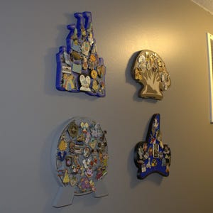 May include: Four decorative pin display boards on a gray wall. The boards are shaped like a castle, a tree, a globe, and a star, each filled with colorful pins. The castle and star are blue, the tree is tan, and the globe is gray.