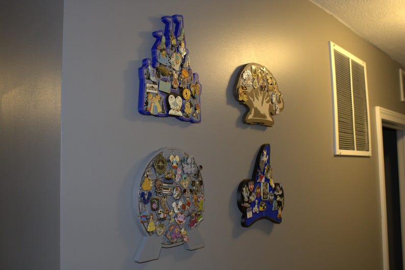 May include: Four decorative pin display boards on a gray wall. The boards are shaped like a castle, a tree, a globe, and a star, each filled with colorful pins. The castle and star are blue, the tree is tan, and the globe is gray.