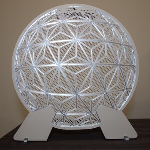 May include: A white, geometric, decorative object with a spherical shape. The design features a repeating pattern of interconnected triangles and star shapes. It stands on two white, rectangular supports.