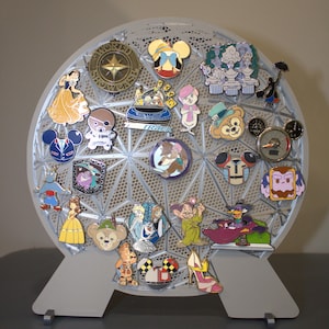 May include: A collection of enamel pins featuring various cartoon characters and designs, displayed on a gray, geometric pin holder. The pins showcase a variety of colors and shapes, including Mickey Mouse, princesses, and other themed characters.