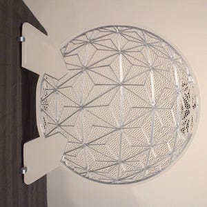 May include: A white, geometric-patterned decorative wall art piece. The art is a semi-circle with a complex design of interconnected shapes and small perforations. It is mounted on a dark wooden surface with two white, rectangular mounting brackets.