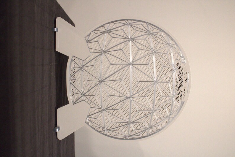 May include: A white, geometric-patterned decorative wall art piece. The art is a semi-circle with a complex design of interconnected shapes and small perforations. It is mounted on a dark wooden surface with two white, rectangular mounting brackets.