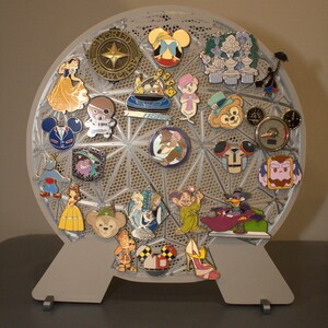 May include: A gray, geometric pin display stand holding a collection of Disney character pins. The pins feature characters like Mickey Mouse, Snow White, and others. The stand is on a gray surface.