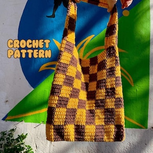 May include: A handmade crochet tote bag with a checkerboard pattern in shades of brown and yellow. The bag has long straps and the words "CROCHET PATTERN" are in the upper left corner. The bag is held up against a wall with colorful graffiti.