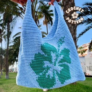 Può includere: Una borsa tote in crochet blu chiaro con un design a fiore di ibisco verde. La borsa ha una tracolla lunga ed è perfetta per portare con sé gli oggetti essenziali di tutti i giorni.