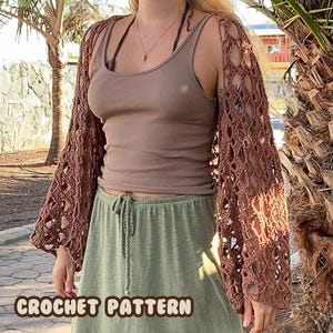 May include: A brown crochet top with a lacy pattern. The top has long sleeves and a loose fit. It is worn over a brown tank top and a green skirt.