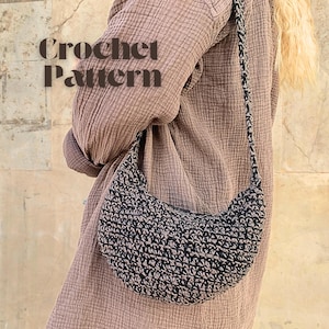Op de afbeelding: Een zwarte en beige gehaakte halvemaanvormige tas met een lange riem. De tas wordt over de schouder gedragen. De tekst "Crochet Pattern" wordt in bruine letters weergegeven.