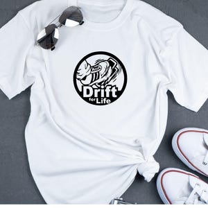 May include: White t-shirt with a black circular graphic featuring a stylized car and the words "Drift for Life." The shirt is paired with sunglasses and white sneakers.