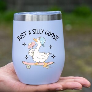 May include: A pale blue insulated tumbler with a silver rim. The tumbler has a cartoon goose on a skateboard, holding a flower, and the text "JUST A SILLY GOOSE". The design includes stars.