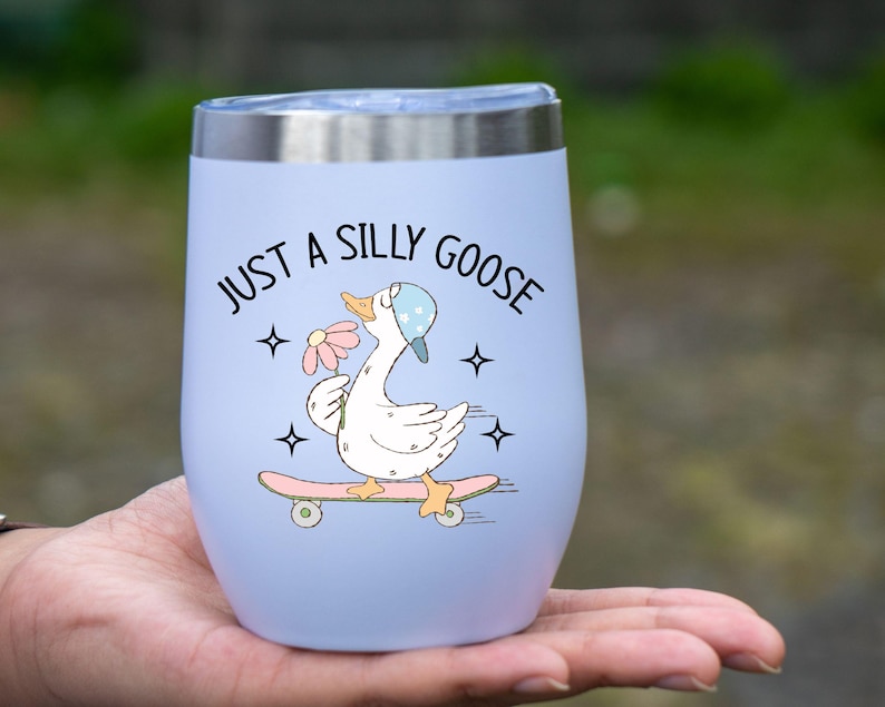 May include: A pale blue insulated tumbler with a silver rim. The tumbler has a cartoon goose on a skateboard, holding a flower, and the text "JUST A SILLY GOOSE". The design includes stars.