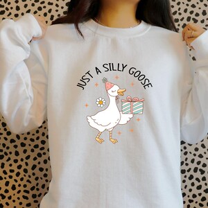 May include: White sweatshirt with a cartoon goose wearing a party hat and holding a gift. The text "JUST A SILLY GOOSE" is arched above the goose. The goose is white with yellow feet and a pink beak. A daisy is next to the goose.