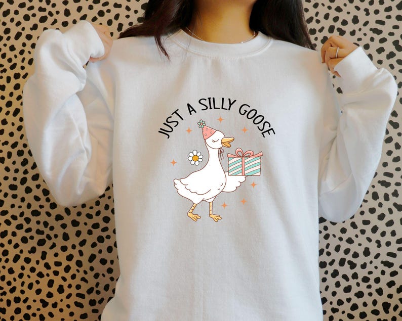 May include: White sweatshirt with a cartoon goose wearing a party hat and holding a gift. The text "JUST A SILLY GOOSE" is arched above the goose. The goose is white with yellow feet and a pink beak. A daisy is next to the goose.
