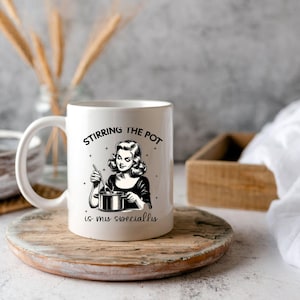 May include: White ceramic mug featuring a black and white illustration of a woman stirring a pot. The text on the mug reads "Stirring the pot is my speciality."