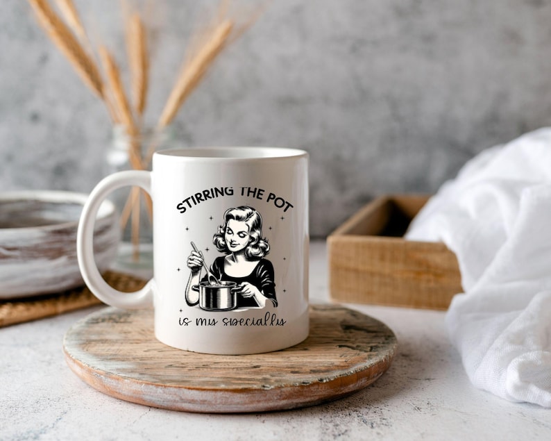 May include: White ceramic mug featuring a black and white illustration of a woman stirring a pot. The text on the mug reads "Stirring the pot is my speciality."