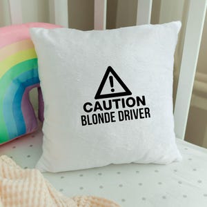 May include: A white, fluffy square pillow with the text "CAUTION BLONDE DRIVER" in black. The pillow has a black triangle with an exclamation point inside. A rainbow-shaped pillow and a pink blanket are in the background.