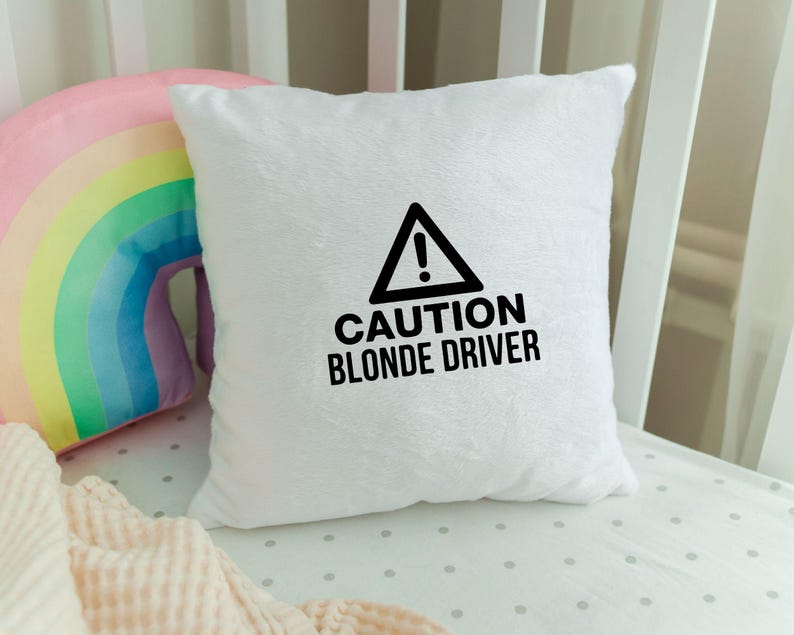 May include: A white, fluffy square pillow with the text "CAUTION BLONDE DRIVER" in black. The pillow has a black triangle with an exclamation point inside. A rainbow-shaped pillow and a pink blanket are in the background.