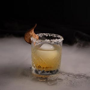 Puede incluir: Un cóctel con un líquido claro y un gran cubito de hielo en un vaso corto y transparente con un borde de sal. Una rodaja de pera seca está en el borde del vaso. La bebida está sobre una superficie blanca con una ligera niebla alrededor.