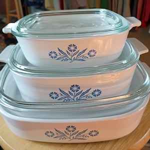 May include: A set of three white rectangular glass baking dishes stacked vertically, each featuring a blue floral pattern on the front. The dishes have integrated side handles and clear glass lids, with the top dish covered by a lid.