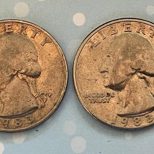 May include: Two silver-colored US quarter dollar coins with the inscription "LIBERTY" on the obverse and "IN GOD WE TRUST" on the reverse. Both coins are dated 1983.