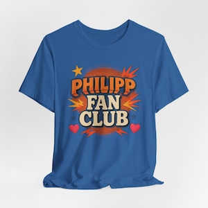 May include: A blue t-shirt featuring the text "PHILIPP FAN CLUB" in orange and white, with a star, hearts, and explosion graphics. The shirt is made of a soft material.