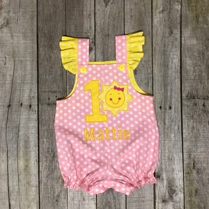May include: Pink and yellow polka dot baby onesie with a yellow sun and the number 1 embroidered on the front. The onesie also has the name Mattie embroidered on the front.