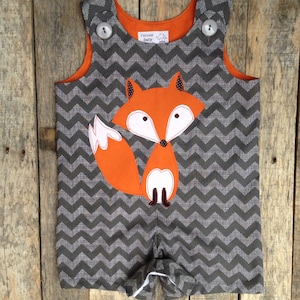 May include: A gray chevron-patterned romper with an orange fox applique. The romper has orange trim, white buttons, and a tag that reads "Fairies Baby". The fox has white accents and black details.