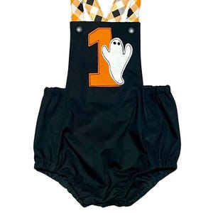 May include: Black Halloween-themed romper with orange number 1 and a white ghost applique. The suspenders feature a plaid pattern in orange, black, and white. The romper has a gathered waist and leg openings.