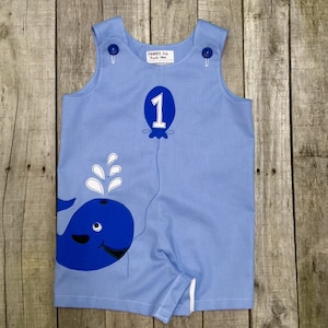 May include: A light blue baby onesie with a blue whale and a blue balloon with a white number 1. The onesie has a white label that says "PAMES Baby" and "Made 18m".