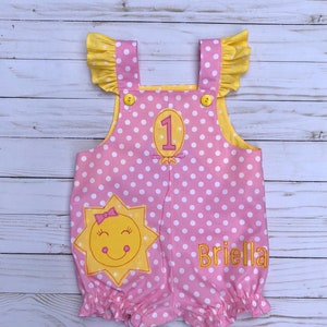 May include: Pink and yellow polka dot romper with a smiling sun and the number 1 on the front. The romper has yellow ruffled sleeves and the name "Briella" is embroidered on the bottom.