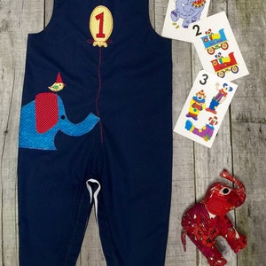 May include: A navy blue overall with a red and blue elephant applique, a yellow balloon with the number "1" on it, and a small bird on top of the elephant. The overall has yellow buttons on the straps. There are three cards in the background with images of a circus elephant, a train, and clowns.