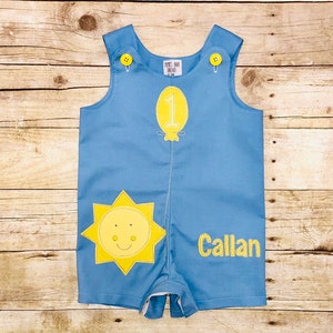 May include: Light blue baby romper with yellow sun and balloon appliqués. Features the number 1 on the balloon and the name "Callan" embroidered in yellow. Includes yellow buttons on the straps.