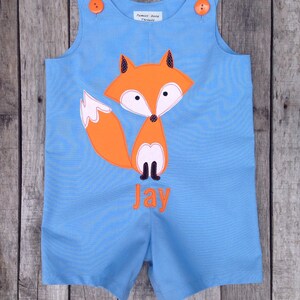 May include: Blue overalls with a fox applique and the name "Jay" embroidered on the front. The overalls have orange buttons and are made by "Pamper Baby Treasures".