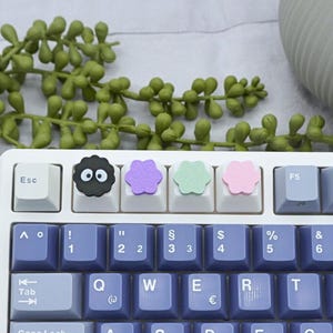 Könnte beinhalten: Eine mechanische Tastatur mit weißem Rahmen und violetten Tastenkappen im QWERTZ-Layout. Die oberste Reihe enthält vier dekorative Artisan-Tastenkappen: einen schwarzen Rußmännchen-Geist, eine violette Blume, eine hellgrüne Blume und eine hellrosa Blume.