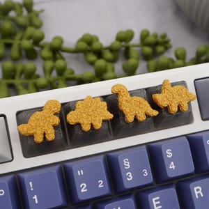 May include: A white mechanical keyboard with blue keys and four gold dinosaur keycaps. The keyboard has an Esc key and function keys. Green leafy sprigs are in the background.