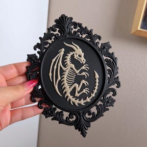 May include: An oval-shaped black frame with intricate scrollwork and floral detailing surrounding a central black disc. A cream-coloured relief sculpture of a dragon skeleton is positioned in the centre of the disc, featuring wings, a tail, and horns.