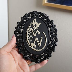 May include: An oval wall plaque featuring a black ornate frame with a matte black centre. A cream-coloured, raised relief illustration of a curled cat skeleton is centred on the black background, showcasing the skull, spine, ribs, and tail.