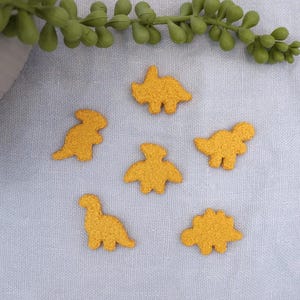 May include: Mustard yellow dinosaur-shaped felt appliques scattered on a light gray fabric. The appliques are in various poses, with textured details. Green plant stems are in the upper left corner.