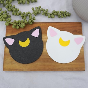 May include: Two cat-shaped coasters, one black and one white, each with pink ears and a yellow crescent moon. They rest on a wooden board, with greenery and a white vase in the background.