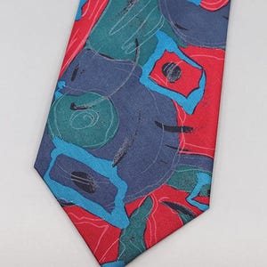Puede incluir: Una corbata estampada con un diseño llamativo. La corbata muestra una mezcla de formas abstractas rojas, turquesas y azul marino. El diseño incluye remolinos, cuadrados y líneas, creando un aspecto audaz y artístico. La corbata es un accesorio clásico.