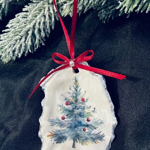 May include: Hand-painted Christmas ornament with a blue Christmas tree and red baubles on a white, shell-shaped base. The ornament is trimmed with silver and has a red ribbon for hanging. Festive holiday decor.