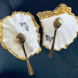 May include: Two white oyster shell-shaped dishes with gold-painted edges, each holding a small gold spoon. One dish features a butterfly illustration. The dishes are set against a dark background, creating a striking contrast.