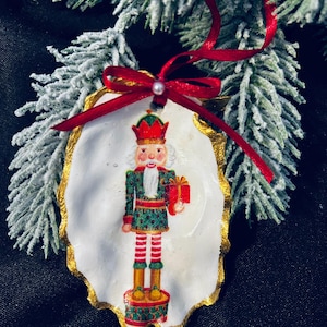 May include: A Christmas ornament with a painted nutcracker design on a white, oyster shell-shaped base with a gold trim. It has a red ribbon and a small pearl. The background includes frosted evergreen branches.