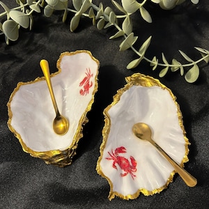 May include: Two oyster shell-shaped dishes with gold rims and small gold spoons. Each white dish features a red crab design. The dishes are on a dark surface, with green foliage in the background.