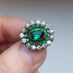 May include: A vintage-style brooch featuring a central oval emerald-green gemstone surrounded by smaller green and clear, sparkling crystals. The brooch has a silver-colored metal setting, creating a classic and elegant design.